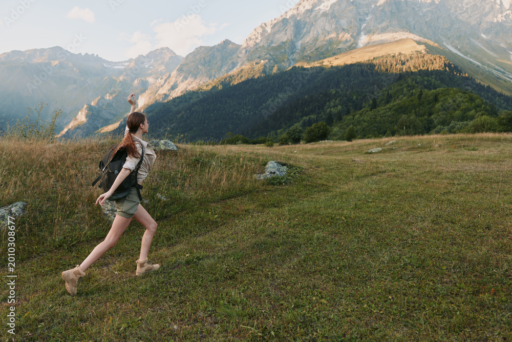 Naklejka premium Young woman hiking in a grassy mountain meadow, exploring nature under blue skies, carrying a light backpack, and enjoying fresh air. Her casual outdoor outfit and sturdy boots convey a relaxed,