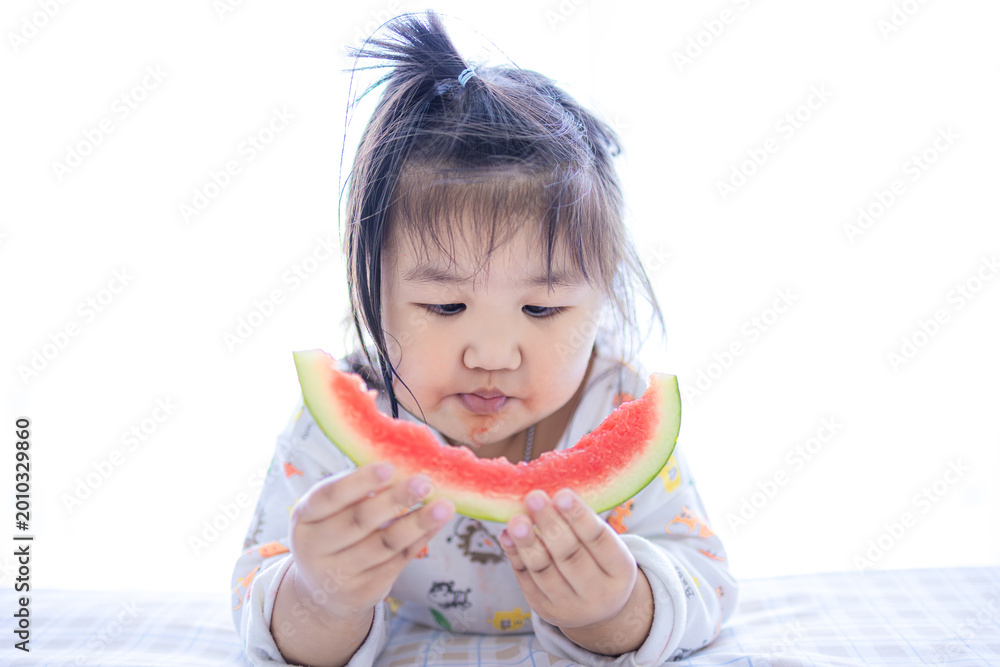 Fototapeta premium Adorable Asian Toddler Girl Eating Juicy Watermelon Slice with Enjoyment,A cute little Asian girl with a ponytail lying down and holding a half-eaten slice of fresh watermelon. She looks focused.
