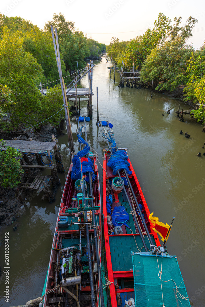 Fototapeta premium Two Traditional Thai Longtail Fishing Boats Moored in a Mangrove Canal at Sunset,High-angle view of two vibrant red and green traditional longtail fishing boats docked side-by-side in a narrow canal.
