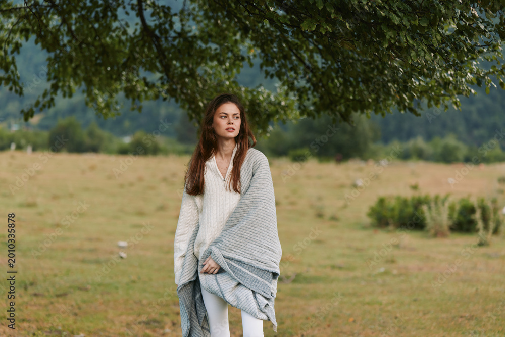 Naklejka premium Woman in a gray shawl stands in an open field, natural lifestyle portrait, calm mood, soft light, outdoor countryside setting, serene and relaxed, she gazes into distance, casual pose, windswept