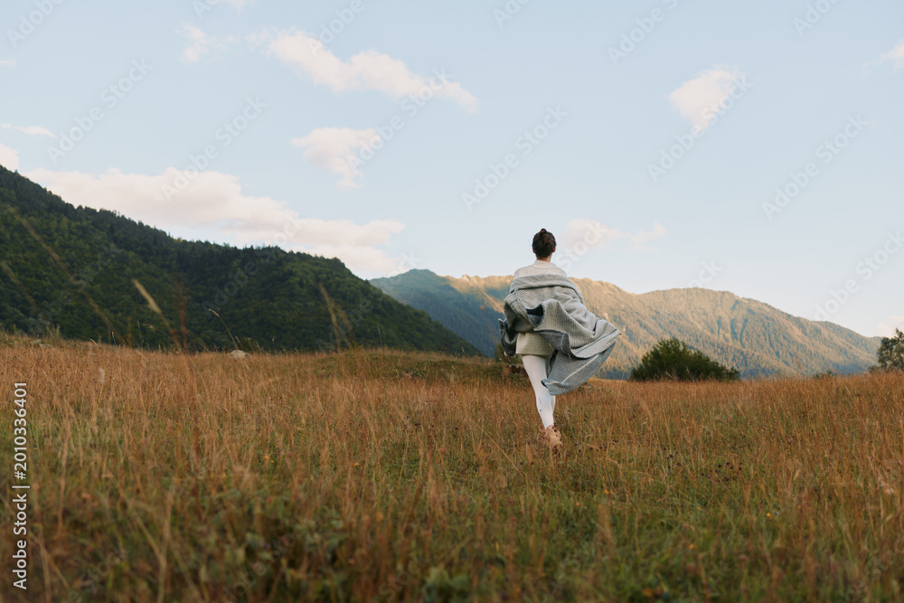 Fototapeta premium Woman walking through a tall grass field toward distant mountains, embracing outdoor living, calm sunlight, free spirit, natural landscape, serene mood, in the golden hour breeze quiet sky above.