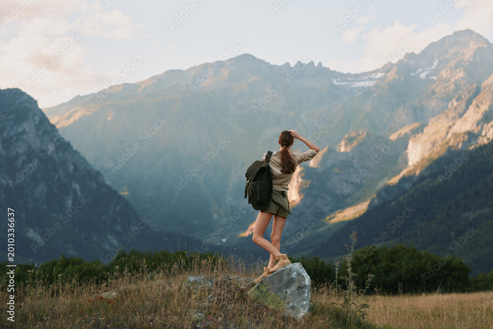 Naklejka premium A young woman with a backpack balances on a sun-warmed rock in a wide mountain meadow, gazing toward distant peaks with a sense of exploration, freedom, and awe.