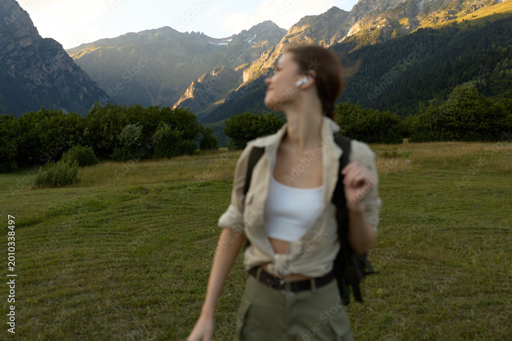 Fototapeta premium A woman in light outdoor attire walks through a grassy meadow with distant mountains, carrying a backpack. Concept for solo travel, nature exploration, and casual lifestyle.