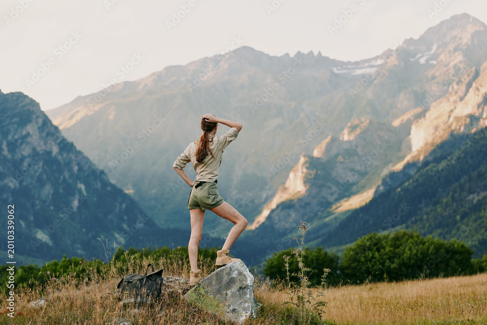 Fototapeta premium A woman stands on a rock in an alpine meadow, looking toward distant peaks, capturing a moment of outdoor adventure, personal exploration, and calm freedom.