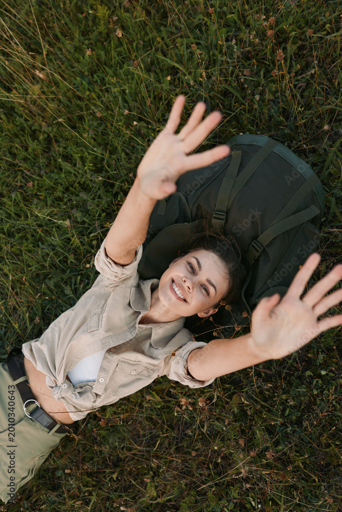 Naklejka premium A smiling woman relaxes on grass outdoors, arms raised in a carefree pose. Casual outfit, natural light, and green surroundings convey a joyful, laidback lifestyle.