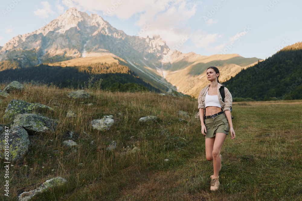 Fototapeta premium A woman hiking in a sunlit field with towering mountains in the distance, capturing outdoors lifestyle, adventure, and nature within a vivid landscape for stock use.