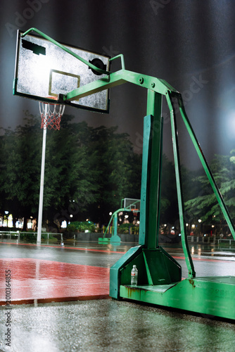 Green Basketball Hoop on Wet Outdoor Court at Night with Trees and Lights in Background