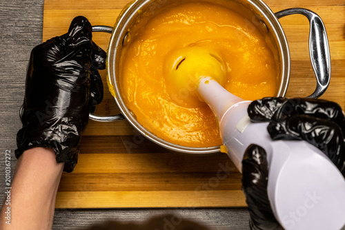 Photos Top view chef in black gloves uses immersion blender to grind and emulsify pumpkin in saucepan in hot state