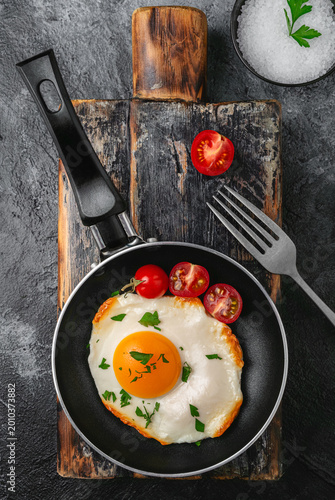 Sunny side up egg in a frying pan with cherry tomatoes and herbs on a rustic wooden board. Dark textured background, top view. Simple homemade breakfast concept.