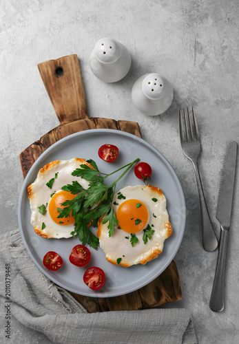 Two sunny side up fried eggs with halves of cherry tomatoes and fresh parsley sprigs on a grey ceramic plate and wooden cutting board, light stone background, top view.