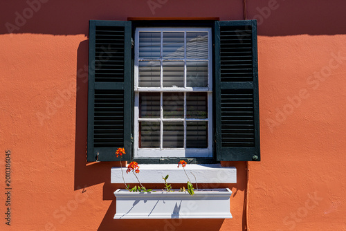 White painted window with black shutters on a bright orange Bermudan house with orange  Epidendrum Orchids in a white window box