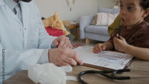 Medium close up handheld shot of small African American boy drawing on medical document while sitting at table with mother and male doctor during home visit