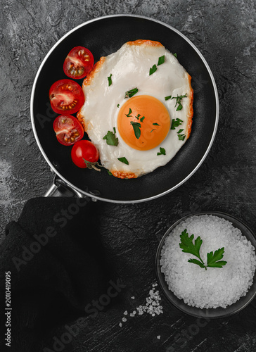 Fried egg with vibrant yolk in a small black skillet with a kitchen towel on the handle, served with cherry tomatoes and sea salt on a dark stone background, top view.