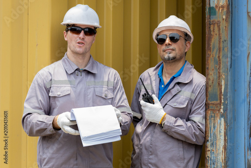 Tableau sur toile Two male construction workers in safety gear stand by a container, holding documents and a walkie-talkie, ready for site coordination