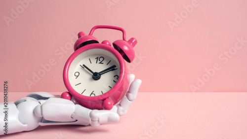 Fotografie Description: White robotic hand holding a pink vintage alarm clock against a pastel pink background representing AI time management and future technology