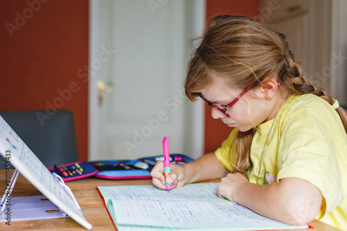 Cute elementary school girl writing in her workbook and doing school homework. Focused child learning and studying indoors. Concept of education, childhood, home learning.