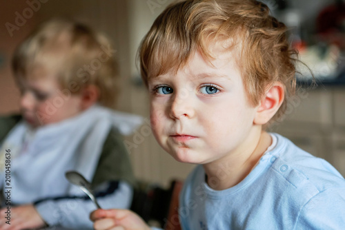 Two little boys eating meal in kitchen at home or daycare nursery, siblings enjoying food together at table, candid family lifestyle moment, real life, real people.