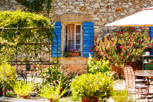 Sunny courtyard in Provence France with stone house, blue shutters and blooming flowers, cozy garden terrace, charming rural scene.
