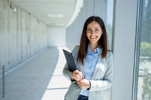 Smiling businesswoman holding clipboard standing in modern office corridor
