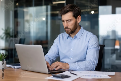 Focused businessman working on laptop in modern office, professional male employee typing on computer, serious man concentrating on business tasks, dedicated worker in corporate environment.