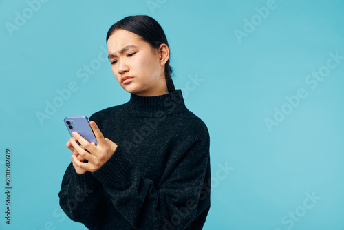 Woman in black sweater looking confused or frustrated at smartphone with blue background. Expression of doubt or concern while checking mobile device in casual studio setting.