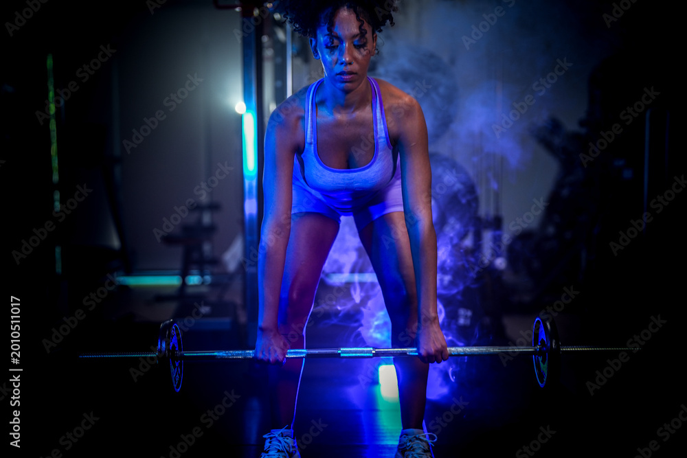 Naklejka premium A woman in purple sportswear preparing for a heavy lift with a barbell in a moody, low-light gym environment with dramatic highlights.