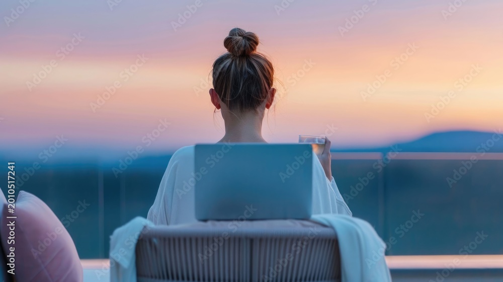Obraz premium View from behind of a woman relaxing on a balcony with her laptop and a drink enjoying a vibrant sunset