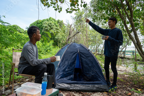Two man tourists backpacking and relaxing on picnic in forest with camping equipment. Man pitching tent on camping trip. Vacation concept on a sunny summer day.