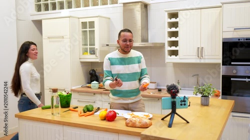 Young man vlogging about cooking healthy food while his girlfriend surprisingly joins him, hugging, dancing, and laughing together in front of the smartphone camera on a tripod for a live stream