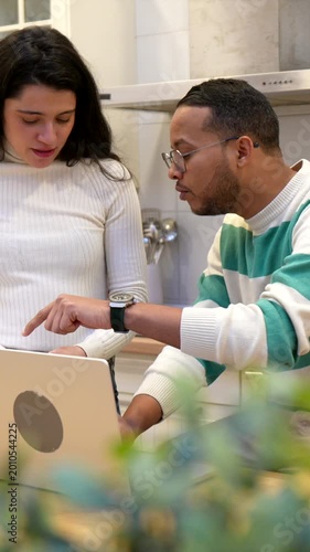 Happy diverse couple reviewing documents and using a laptop in their kitchen, managing household finances, paying bills online, and planning their budget with a positive and collaborative attitude