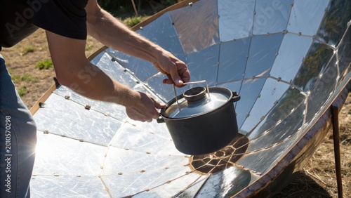 A person uses a solar cooker with reflective panels to heat a pot outdoors in a sunny environment.