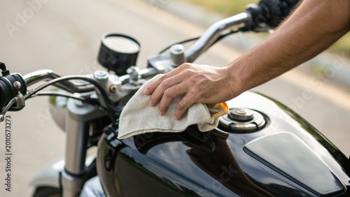 A person is cleaning a black motorcycle fuel tank with a cloth, focusing on maintenance and care of the bike in an outdoor setting.