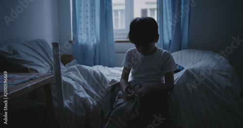 Boy sitting on bed in dark bedroom with head down, conveying sadness, melancholy, and signs of childhood depression