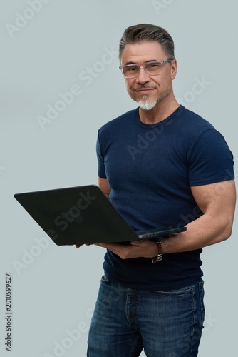 Casual mid adult man standing holding laptop computer. Portrait of happy middle aged male in 50s with gray hair and glasses, smiling. Isolated on white background.