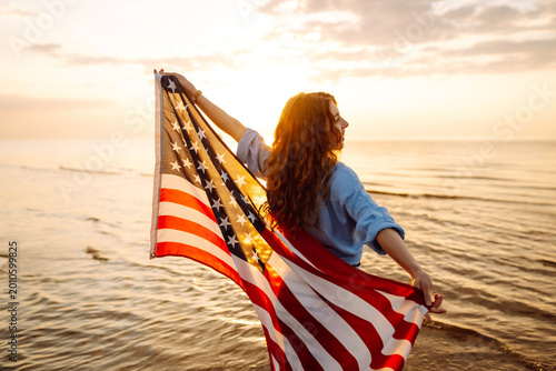 Slika na platnu A cheerful woman stands on the seashore with an American flag at sunset