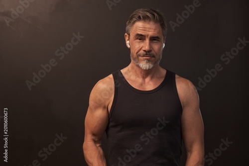 Portrait of happy muscular athletic mid adult man standing against dark background, smiling. Studio shot of middle aged confident sporty male fitness trainer with gray hair.
