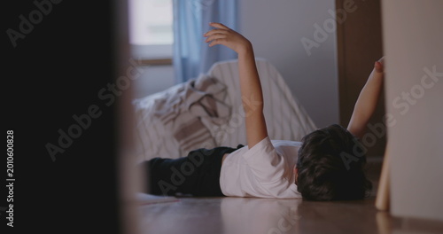 Boy lying on bedroom floor with arm raised in air, appearing bored and restless, symbolizing childhood idleness, lack of engagement, and waiting in silence indoors