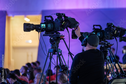 Cameraman operates professional video camera on tripod during live conference, colorful stage lighting and audience.