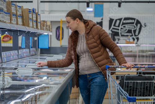 Fotografi Woman Checking Prices In Supermarket Aisle, Comparing Brands And Scanning Tags, Practical Posture With Cart, Everyday Outfit, Budgetminded Grocery Run Atmosphere With Visible Shelf Signage