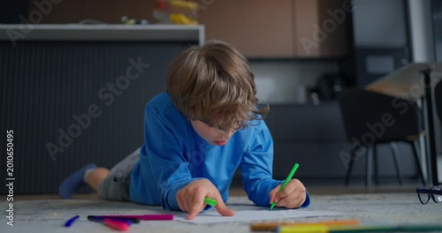 Young boy lying on carpeted floor drawing with colorful markers in a modern home interior, focused on creative play and childhood learning