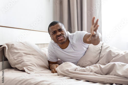 Valokuva Frustrated African American man lying in bed, reaching arm out with irritated expression as if dismissing or gesturing