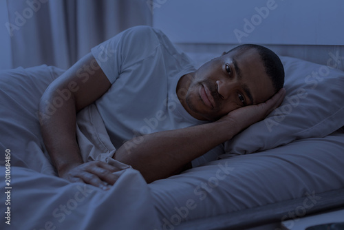 Fotografija Sleepless African American man in white t-shirt lying in bed at night, eyes open looking at camera with a tired expression in dark blue light