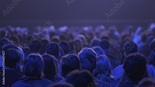Large audience seated in dark theater under blue stage lighting, focused attention, shared anticipation, quiet atmosphere.