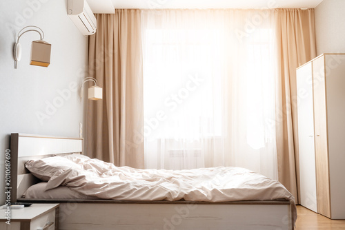 Bright, cozy bedroom interior with unmade bed, beige curtains, wall lamp, wardrobe, and air conditioner. Warm morning sunlight floods the minimalist room