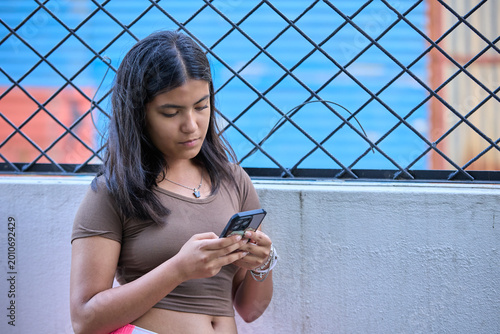 Young woman in brown top standing outdoors near chain-link fence focused on smartphone