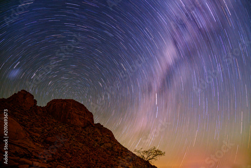 Star trails over Spitzkoppe granite inselberg, Namibia, long exposure night sky, circular star rotation, Milky Way, lone tree silhouette, desert landscape, astrophotography, Africa