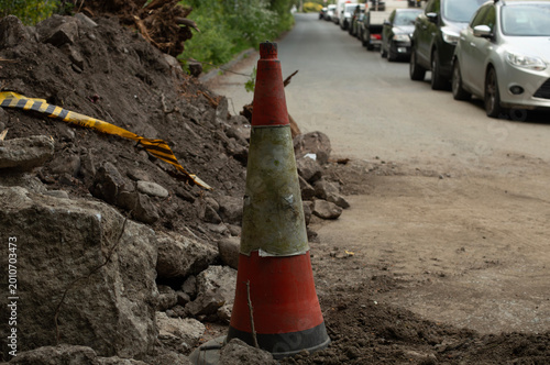 A weathered traffic cone stands among rocks and loose soil at a roadside construction site. The bright orange and white cone contrasts with the rugged surroundings, serving as a warning marker 