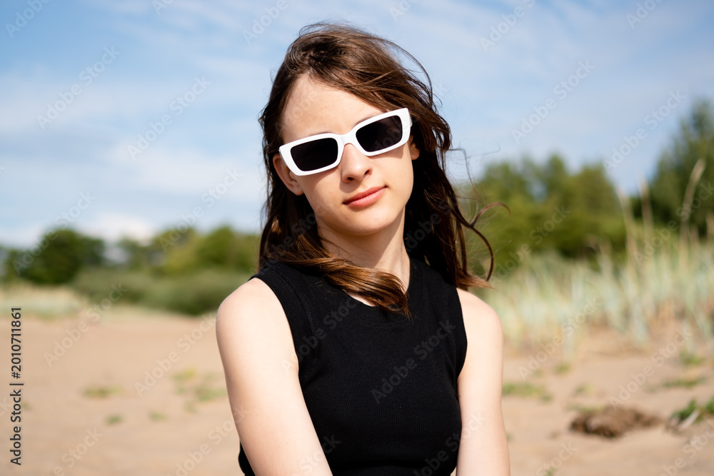 Naklejka premium A teenage girl poses on a sandy field with grass and trees behind, her calm gaze meeting the camera. The teenager radiates natural beauty and relaxed poise in this peaceful outdoor portrait.