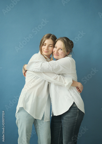 Beautiful mature mother and adult daughter in white shirts and jeans hugging in the photostudio on blue paper background. Two female generations. Mother's day concept