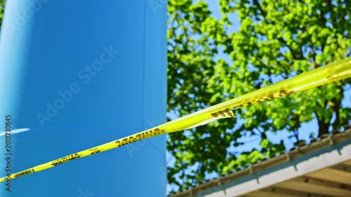 Caution tape is tied around a blue pole on a sunny day. Trees with green leaves are seen in the background. The area is marked for warning, keeping people away.